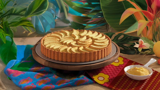 Ein gebackener Apfelkuchen steht auf einem Holzständer, umgeben von tropischen Blättern und Blumen, mit einem bunten Tuch und einer kleinen Schale mit Pudding in der Nähe.
